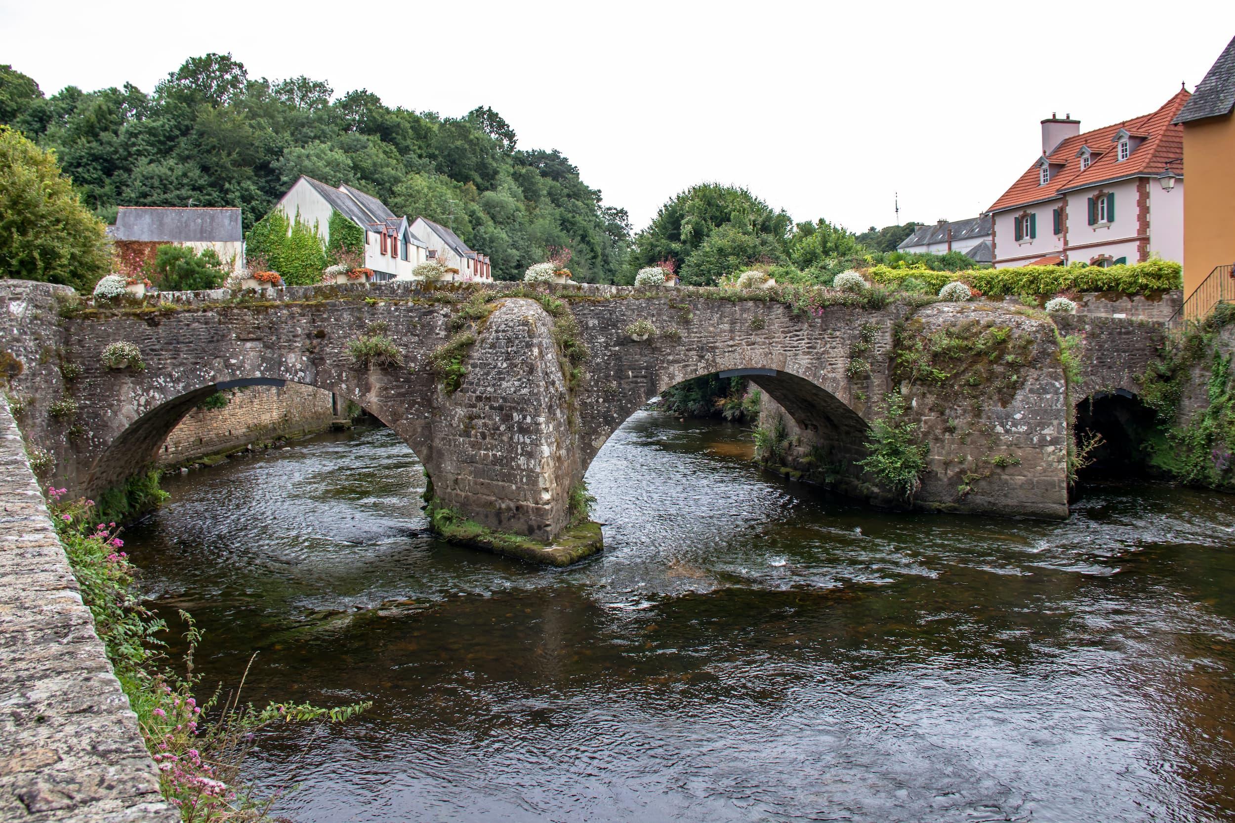 Vieux pont en pierre traversant une rivière dans un village, entouré de maisons traditionnelles et de verdure
