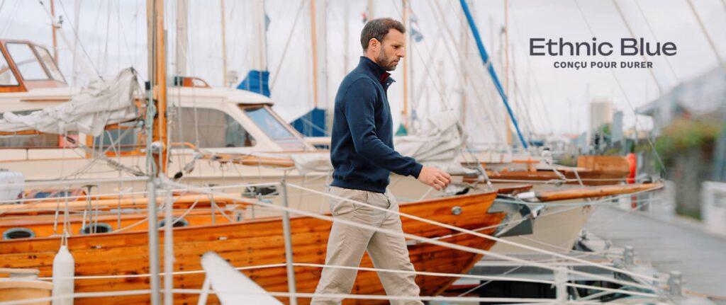 Un homme marche le long d'un ponton bordé de voiliers en bois dans un port. Il porte un pull bleu marine et un pantalon beige, reflétant une allure décontractée et élégante. Le texte 'Ethnic Blue, conçu pour durer' est visible en arrière-plan.