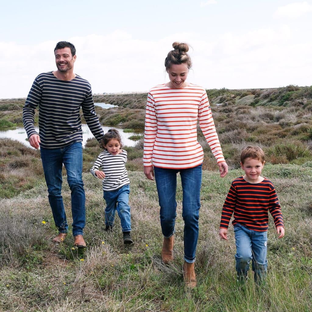 Une famille de quatre personnes, vêtue de marinières assorties, marche joyeusement dans une prairie. Les parents et les deux enfants partagent un moment complice dans un cadre naturel, avec un ciel dégagé en arrière-plan.